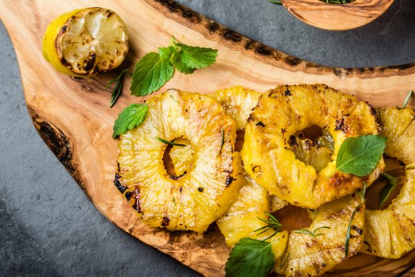 Grilled pineapple slices with fresh mint on olive wooden cutting board, gray slate background. top view