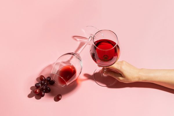Female hand holding a glass with red wine and a bunch of grapes on a gentle pink background. Selective focus. Copy space. Minimalism. Top view flat layout